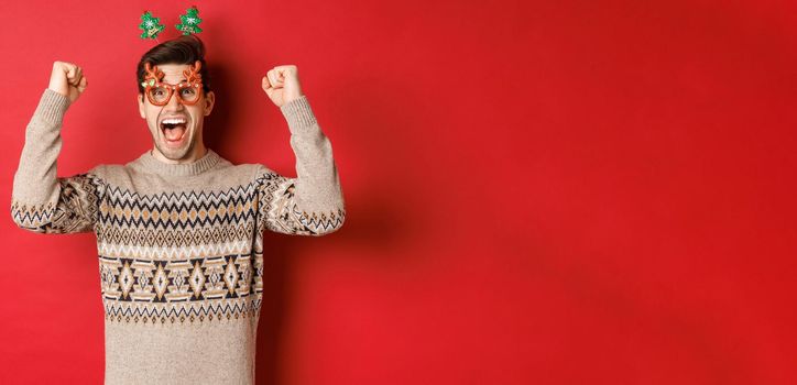 Portrait of thrilled and happy man in party glasses, winter sweater, shouting for joy and raising hands up, winning prize on christmas, achieve new year goal, standing red background.