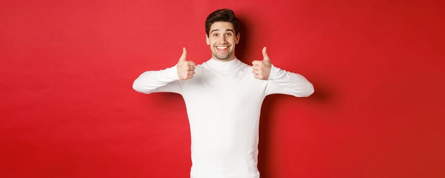 Cheerful good-looking male model in white sweater, showing thumbs-up in approval, like something good, standing over red background and smiling pleased.