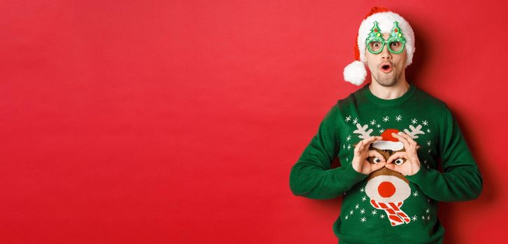 Portrait of carefree handsome man in santa hat and party glasses, making fun of his christmas sweater, looking happy over red background.