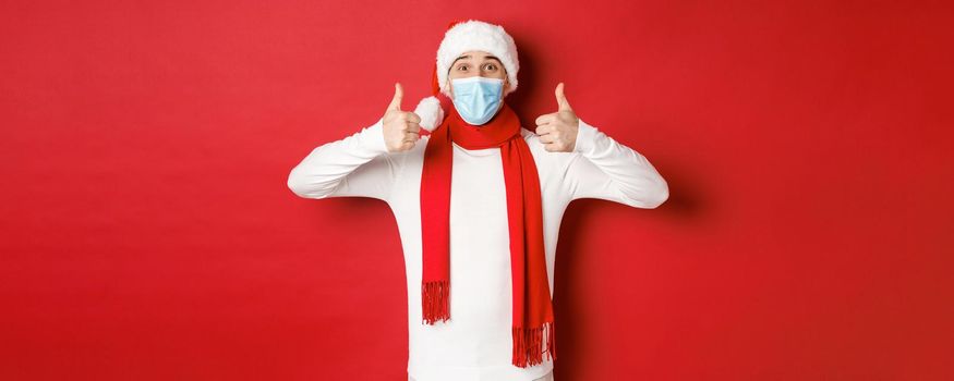 Concept of new year, coronavirus and holidays. Cheerful man celebrating new year and social distancing, wearing medical mask, santa hat and scarf, showing thumbs-up in approval.