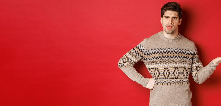 Image of disappointed and confused handsome man, wearing christmas sweater, raising hand and frowning, asking you to explain something, standing over red background.