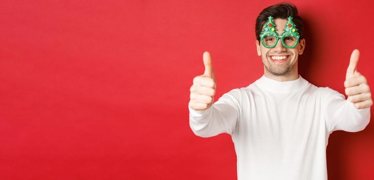 Close-up of attractive happy man in party glasses and white sweater, showing thumbs-up in approval and smiling, standing over red background.