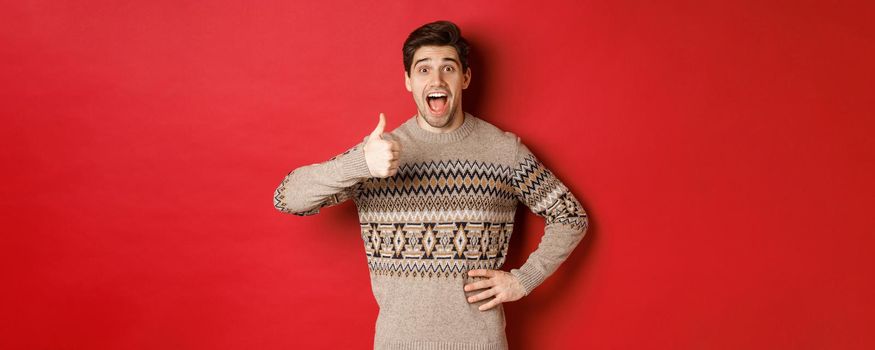 Portrait of excited, attractive man in christmas sweater, showing thumbs-up and looking amazed, recommending good thing, advertising christmas related product.