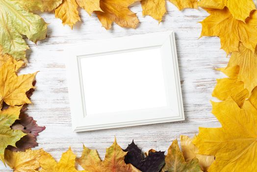 Blank rectangular photo frame lies on vintage wooden desk with bright autumn foliage. Flat lay with autumn leaves on white wooden surface. Simple white picture frame with copy space for design.