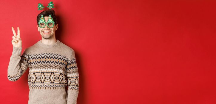 Portrait of funny and cute man in party glasses and christmas sweater, smiling and showing peace sign while posing over red background for new year advertisement.