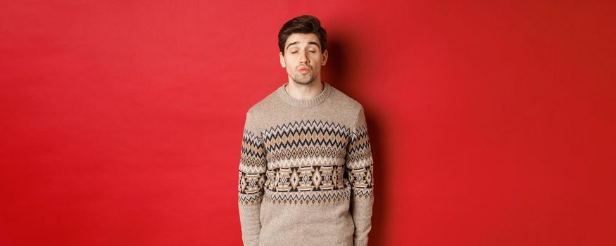 Image of handsome man in christmas sweater pucker lips and close eyes, waiting for kiss under mistletoe, standing over red background.