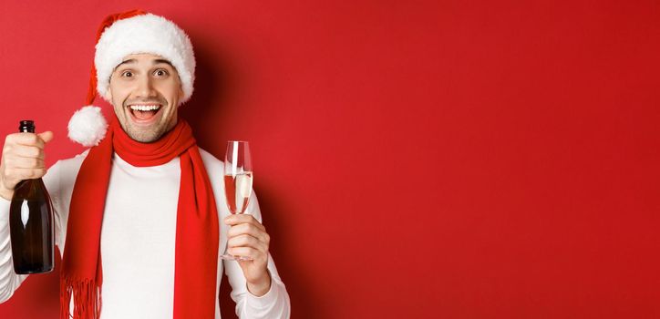 Concept of winter holidays, christmas and lifestyle. Close-up of cheerful handsome man, holding champagne bottle and glass, celebrating new year, standing over red background.