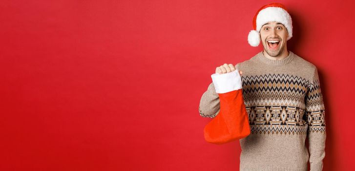 Concept of winter holidays, new year and celebration. Cheerful and surprised adult man receiving candies on Saint Nicholas day in red stocking, standing amazed over red background.