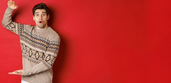 Image of attractive man in christmas sweater, shaping big new year gift, showing something large and amazing, standing over red background.