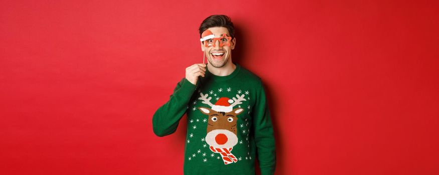 Portrait of handsome smiling man in christmas sweater and party glasses, looking surprised and happy, celebrating new year and having fun, standing against red background.