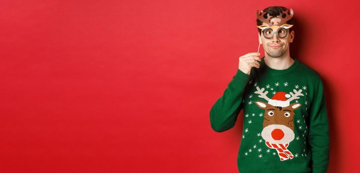 Funny man in christmas sweater and party mask, celebrating winter holidays, showing funny faces, standing against red background.