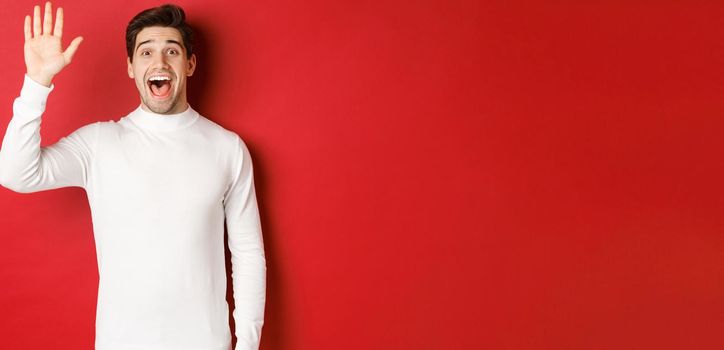 Image of happy and friendly young man saying hello, waving hand to greet someone, standing over red background.