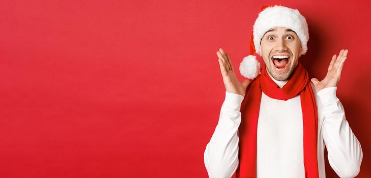 Concept of christmas, winter holidays and celebration. Image of handsome man looking surprised at new year promo offer, smiling amazed, standing over red background.