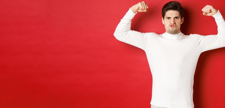 Portrait of handsome and funny guy in white sweater, flex biceps and looking encouraged, showing strong muscles, standing over red background.