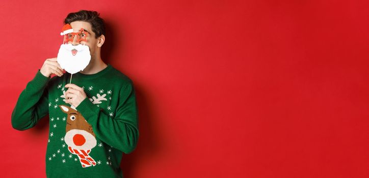 Portrait of surprised man in green christmas sweater, holding funny santa claus mask, celebrating new year, looking left amazed, sanding over red background.