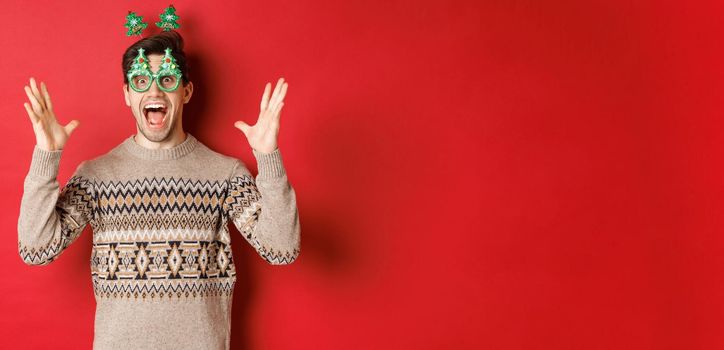 Image of excited and amazed caucasian guy in party glasses, christmas sweater, raising hands up and making big announcement, enjoying new year celebration, red background.