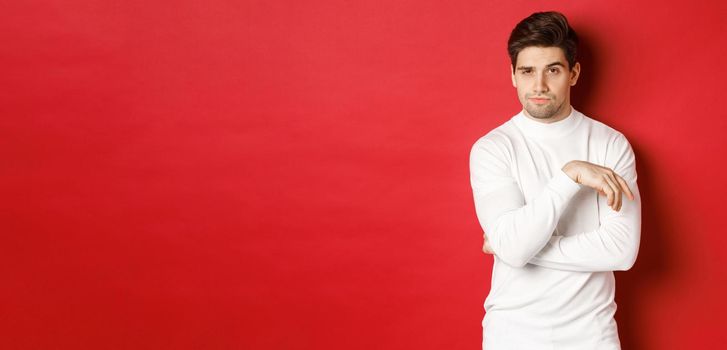 Image of sassy caucasian man in white sweater, looking thoughtful at camera, making choice, standing over red background.