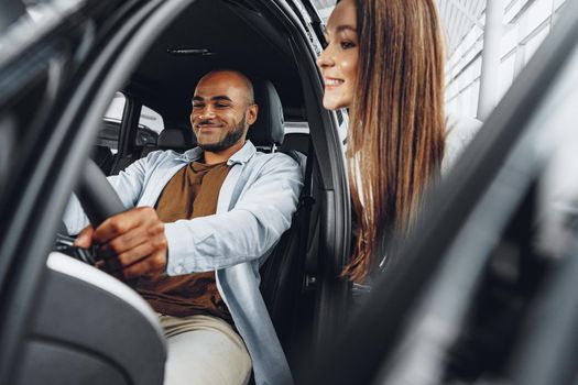 Young attractive caucasian woman salesperson in car showroom showing a car to her male African American client