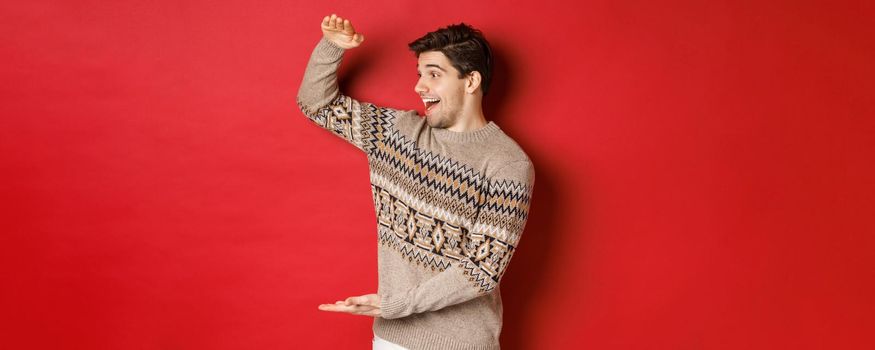 Portrait of happy young man in christmas sweater, showing large present, smiling and looking amazed at cool gift, standing over red background.