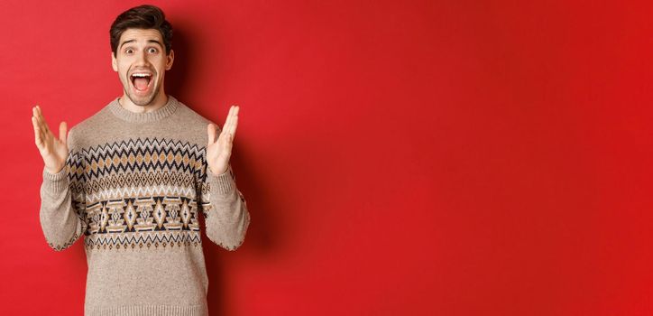 Portrait of surprised and happy handsome man, wearing christmas sweater, looking amazed, celebrating new year, standing over red background.