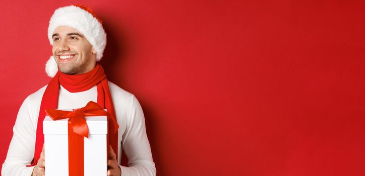 Concept of winter holidays, christmas and lifestyle. Close-up of attractive guy in santa hat and scarf, smiling and looking left while holding new year present, standing over red background.