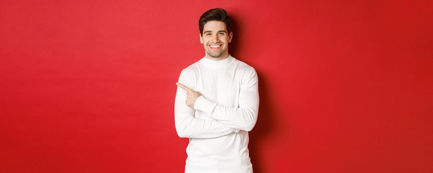 Concept of winter holidays, christmas and lifestyle. Portrait of attractive man in white sweater, pointing finger at upper left corner and smiling, showing logo on red background.