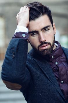 Young bearded man, model of fashion, in urban background wearing british elegant suit. Guy with beard and modern hairstyle touching his hair