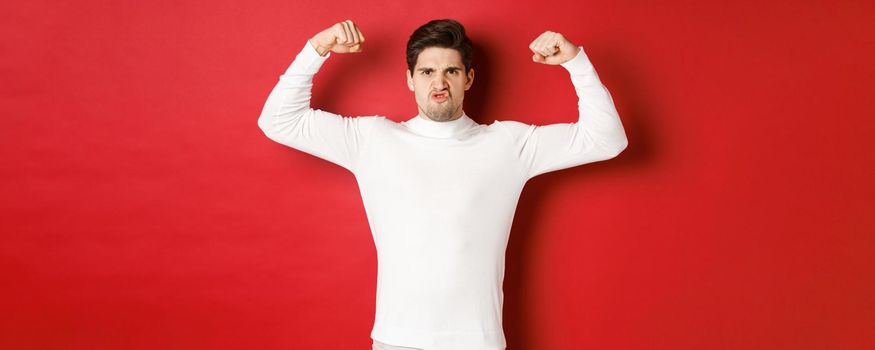 Portrait of handsome and funny guy in white sweater, flex biceps and looking encouraged, showing strong muscles, standing over red background.