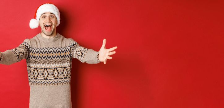 Portrait of happy, attractive man in christmas sweater and santa hat, reaching hands for gift, want to take something and smiling, standing over red background.