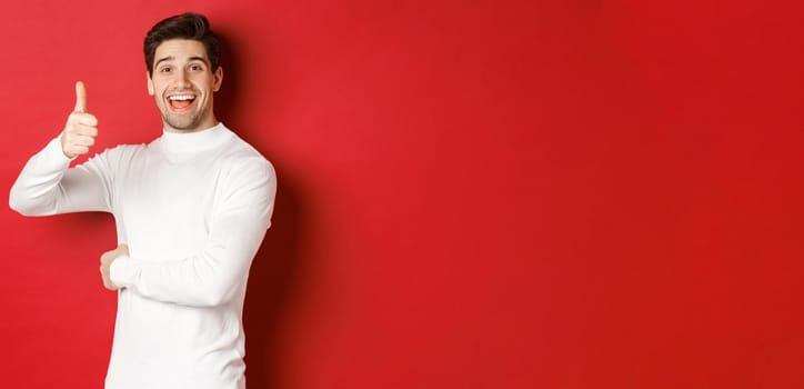 Concept of winter holidays, christmas and lifestyle. Portrait of cheerful, attractive man in white sweater, showing thumbs-up in approval, like good idea, standing over red background.