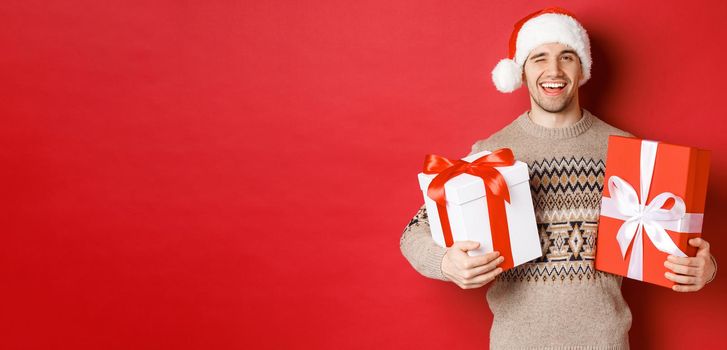 Concept of winter holidays, new year and celebration. Portrait of confident and cheeky young man prepared gifts for christmas, winking and holding presents, standing over red background.