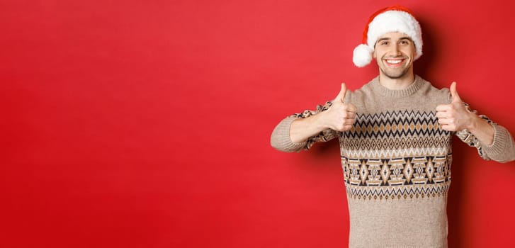 Image of cheerful attractive man in winter sweater and santa hat, showing thumbs-up, celebrating christmas and looking happy, like something, standing over red background.