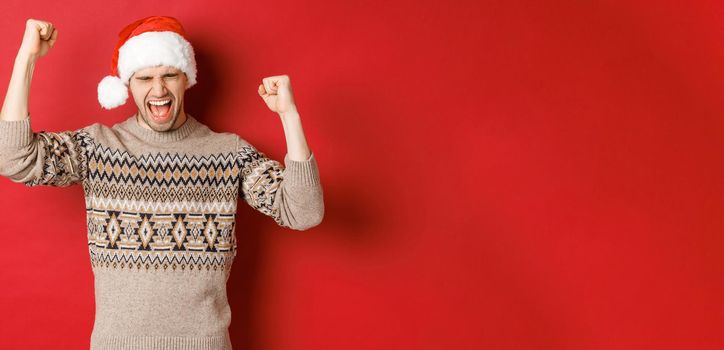 Satisfied handsome man, feeling lucky and happy, shouting for joy and making fist pumps, celebrating victory or win, receive awesome christmas gift, standing in santa hat over red background.