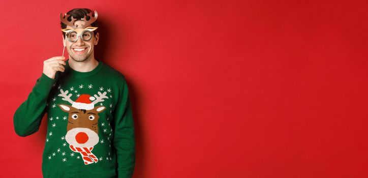 Handsome happy man in christmas sweater, holding party mask and smiling, enjoying new year celebration, standing over red background.