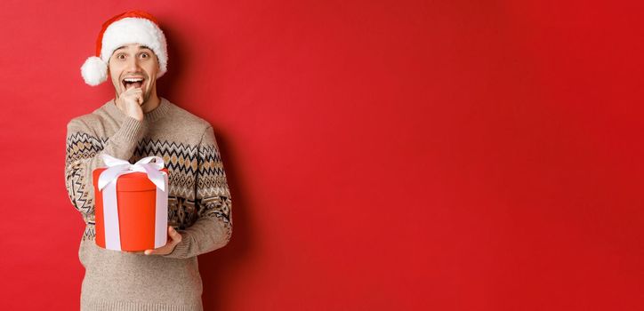 Image of handsome guy in santa hat and christmas sweater, excited to open xmas gift, looking amazed and holding present in one hand, standing over red background.