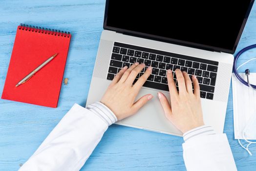 Top view of doctor hands typing at laptop computer. Therapist in white coat sitting at blue wooden desk. Examination and consultation in clinic. Medical application and online healthcare services.