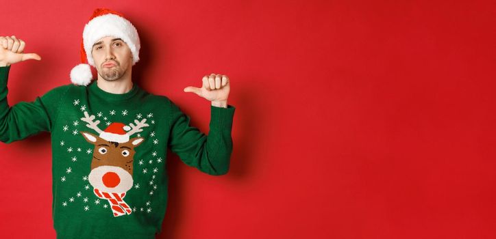 Image of handsome and confident young man in green sweater and santa hat, pointing at himself, celebrating christmas, standing over red background.