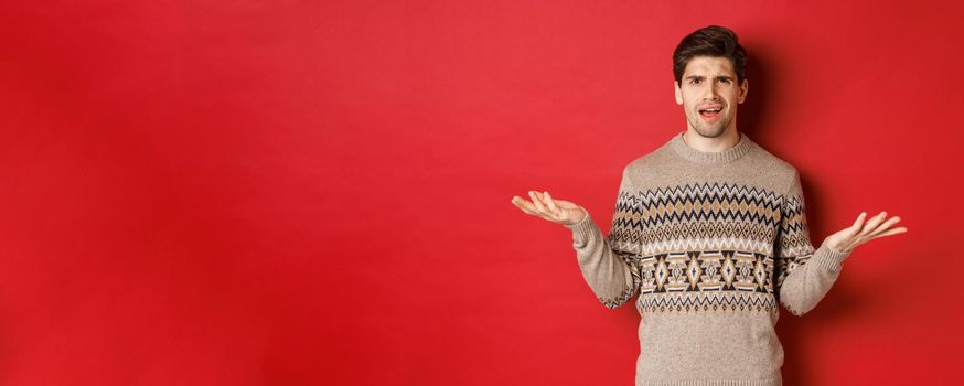 Portrait of confused and disappointed handsome guy, complaining about christmas, spread hands sideways and frowning displeased, standing in xmas sweater over red background.
