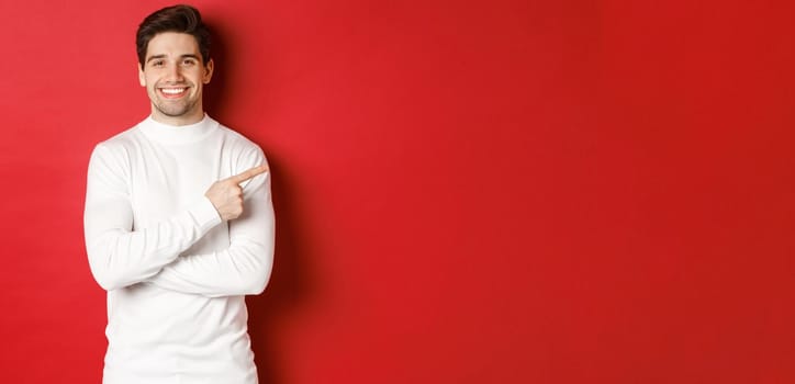 Handsome young man with bristle, wearing white sweater, pointing fingers at upper right corner and smiling, showing christmas banner, standing over red background.