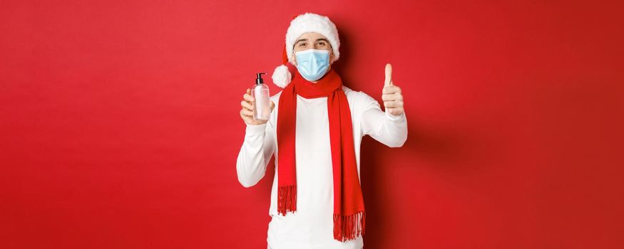 Concept of covid-19, christmas and holidays during pandemic. Happy and satisfied man in santa hat and medical mask, showing thumb-up in approval and recommending hand sanitizer.
