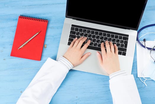 Top view of doctor hands typing at laptop computer. Therapist in white coat sitting at blue wooden desk. Examination and consultation in clinic. Medical application and online healthcare services.