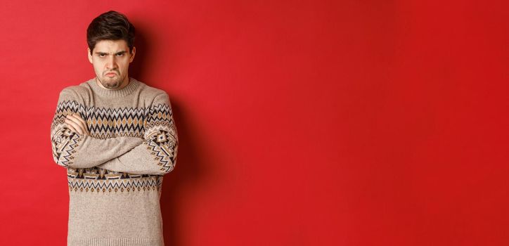 Image of angry and offedned guy in christmas sweater, feeling mad, cross arms on chest and sulking, standing over red background.