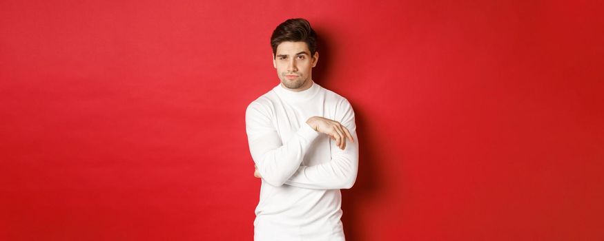 Image of sassy caucasian man in white sweater, looking thoughtful at camera, making choice, standing over red background.