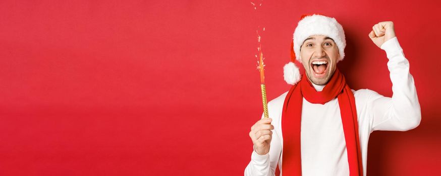 Concept of christmas, winter holidays and celebration. Handsome man celebrating new year and having fun, holding sparkler and smiling, wearing santa hat, standing over red background.