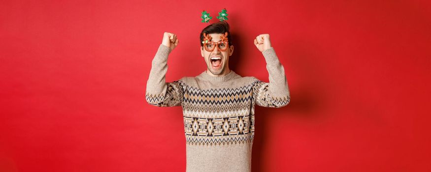 Portrait of thrilled and happy man in party glasses, winter sweater, shouting for joy and raising hands up, winning prize on christmas, achieve new year goal, standing red background.