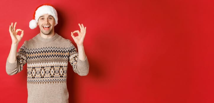 Portrait of happy and pleased handsome man, wearing winter sweater and santa hat, showing okay signs, praise good christmas party, standing over red background.