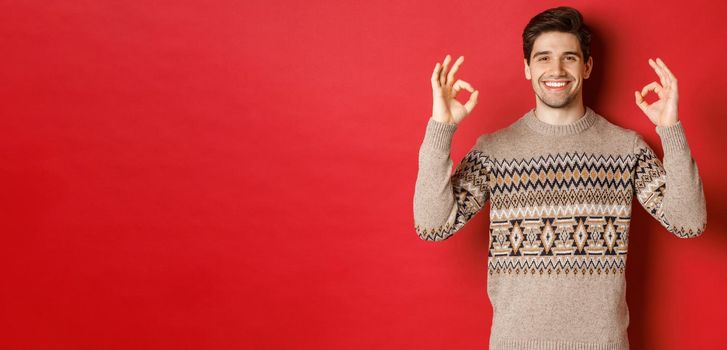 Concept of christmas celebration, winter holidays and lifestyle. Satisfied handsome man in sweater, showing okay signs and smiling pleased, recommending something, red background.