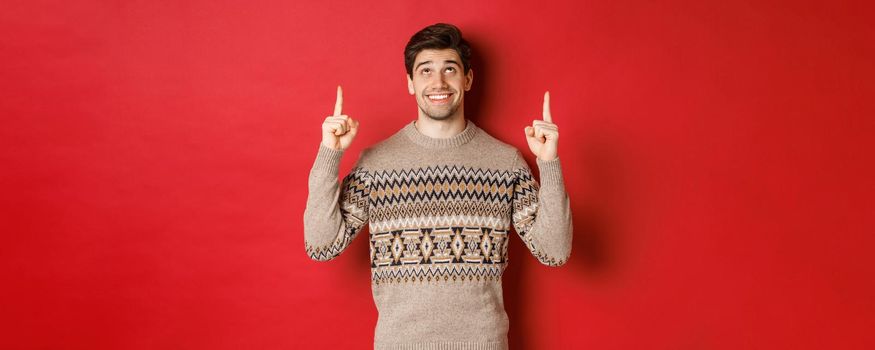 Portrait of dreamy handsome guy thinking about christmas holidays, wearing winter sweater, pointing and looking up at new year promo offer, standing over red background.