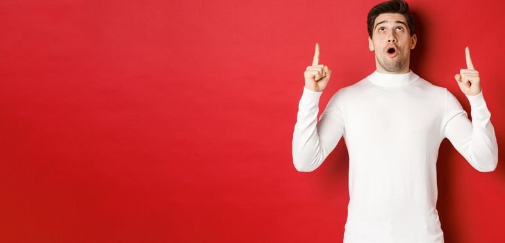 Concept of winter holidays. Portrait of handsome surprised man in white sweater, reacting to new year promo offer, looking and pointing fingers up, standing impressed against red background.