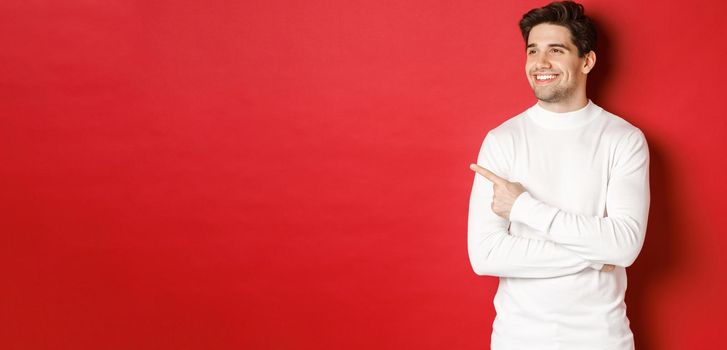 Concept of winter holidays, christmas and lifestyle. Handsome adult man in white sweater, looking and pointing left with pleased smile, showing new year advertisement, red background.